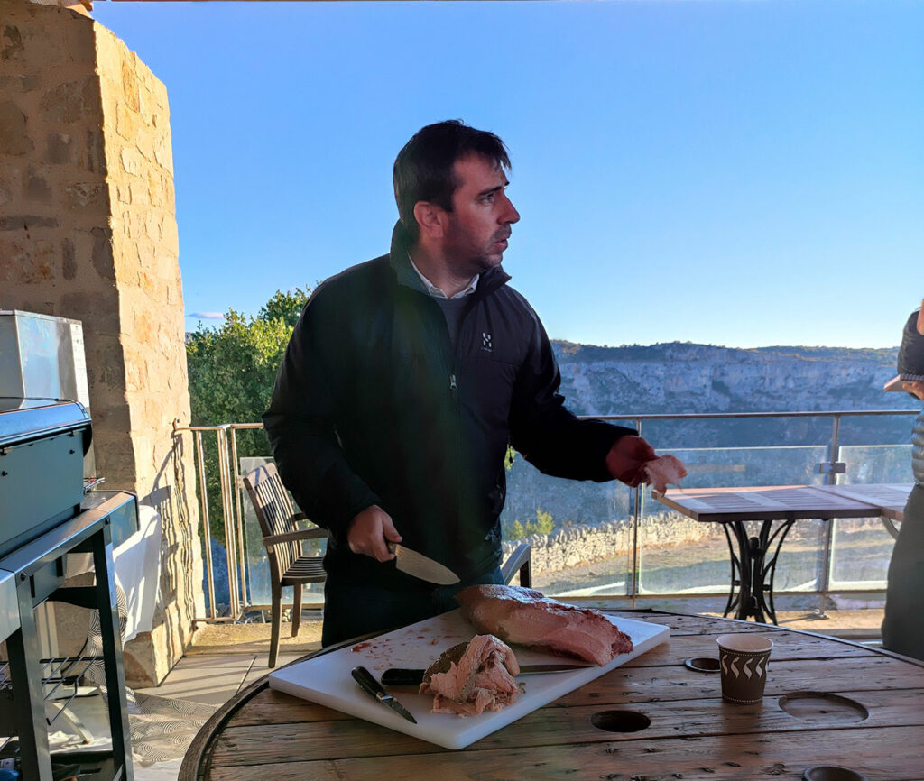 homme brun en train de découper un morceau de viande tendre. En fond se trouve un beau paysage naturel.