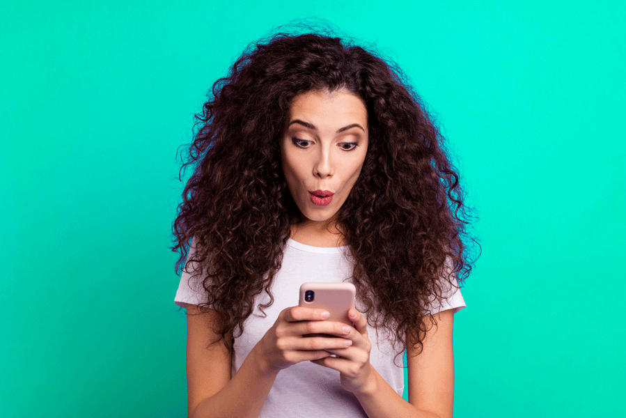 femme aux cheveux bouclés sur un fond vert qui tient sans téléphone entre ses mains et qui a une mine surprise