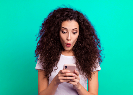 femme aux cheveux bouclés sur un fond vert qui tient sans téléphone entre ses mains et qui a une mine surprise