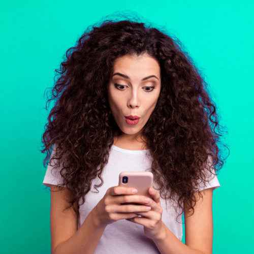 femme aux cheveux bouclés sur un fond vert qui tient sans téléphone entre ses mains et qui a une mine surprise