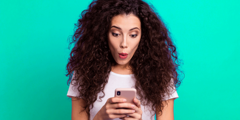 femme aux cheveux bouclés sur un fond vert qui tient sans téléphone entre ses mains et qui a une mine surprise