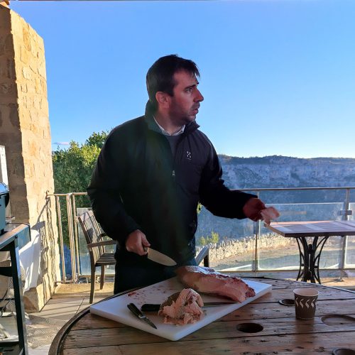 homme brun en train de découper un morceau de viande tendre. En fond se trouve un beau paysage naturel.