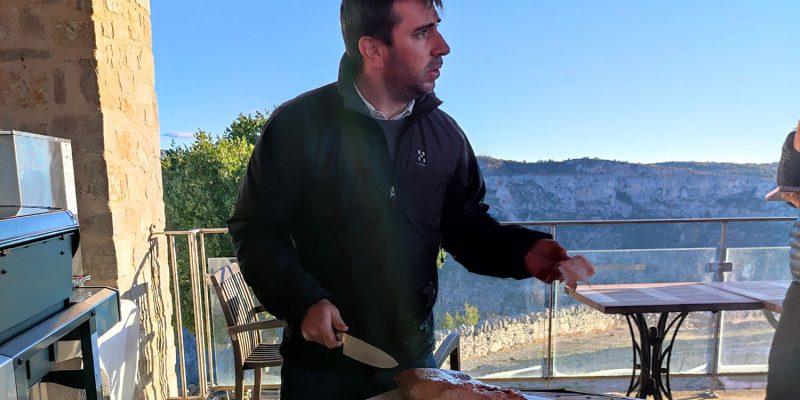 homme brun en train de découper un morceau de viande tendre. En fond se trouve un beau paysage naturel.